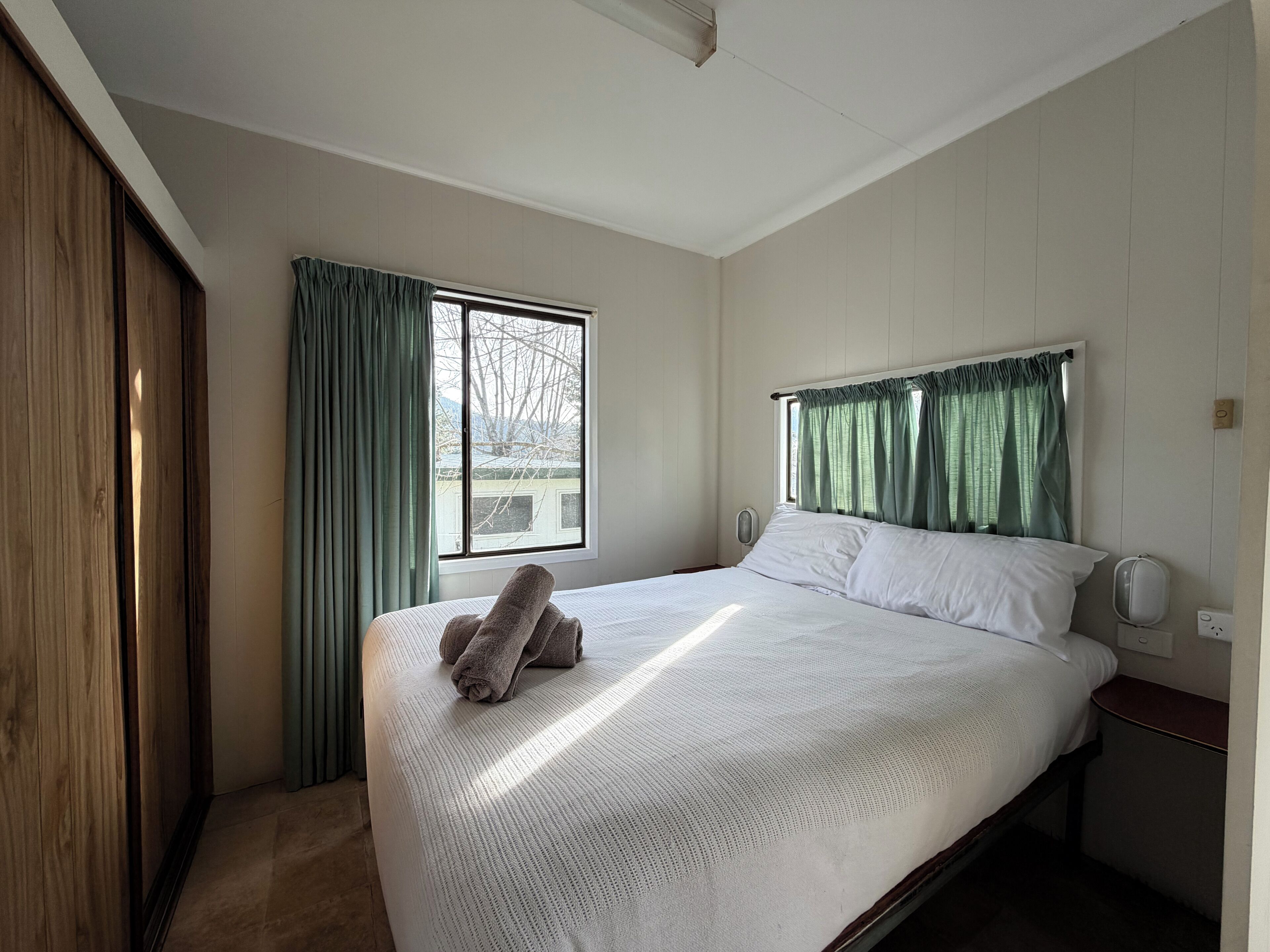 A bedroom with a bed, a window with curtains, a bedside table, and a wooden wardrobe.