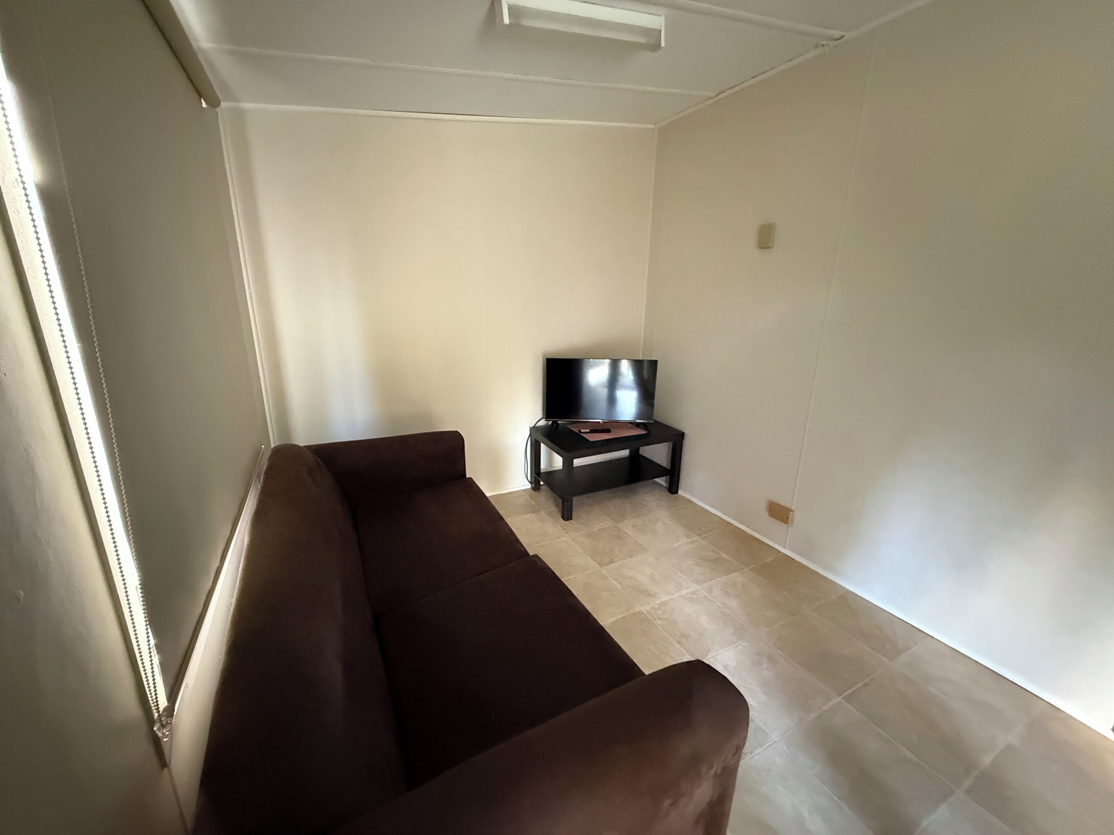 A living room with a brown sofa, a television on a stand, and a tiled floor.