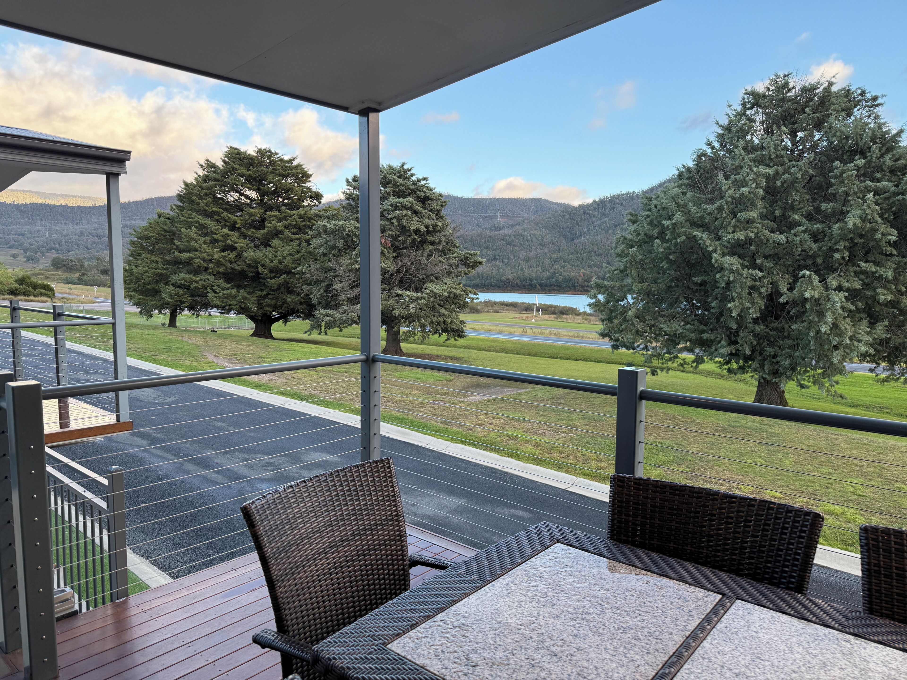 Un espacio de estar al aire libre cubierto con sillas de mimbre y una mesa, con vistas a una carretera y un lago rodeado de árboles y montañas al fondo.