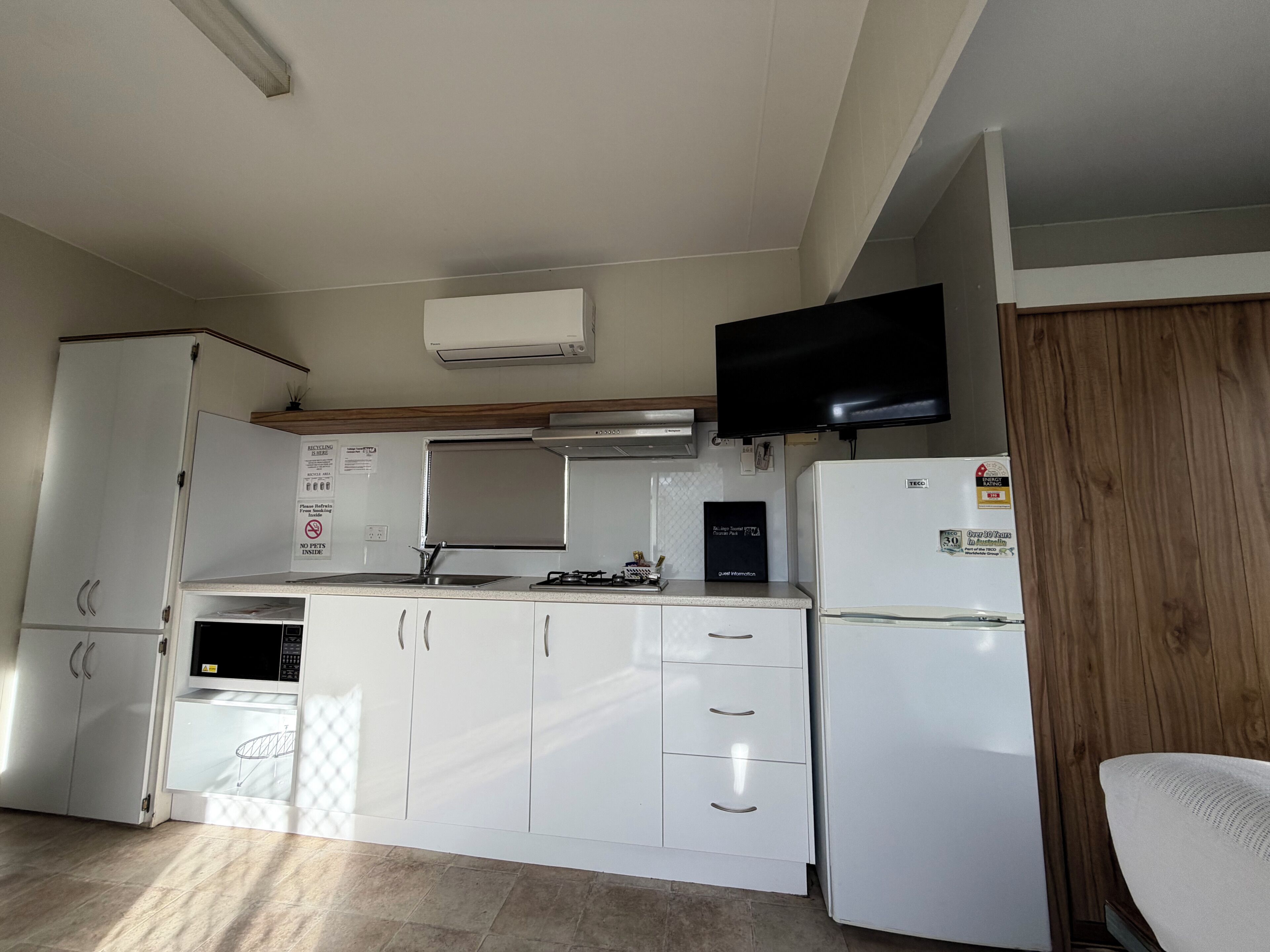 A compact kitchen with white cabinetry, a built-in microwave, a gas stove, a refrigerator, and a flat-screen TV mounted above the stove.