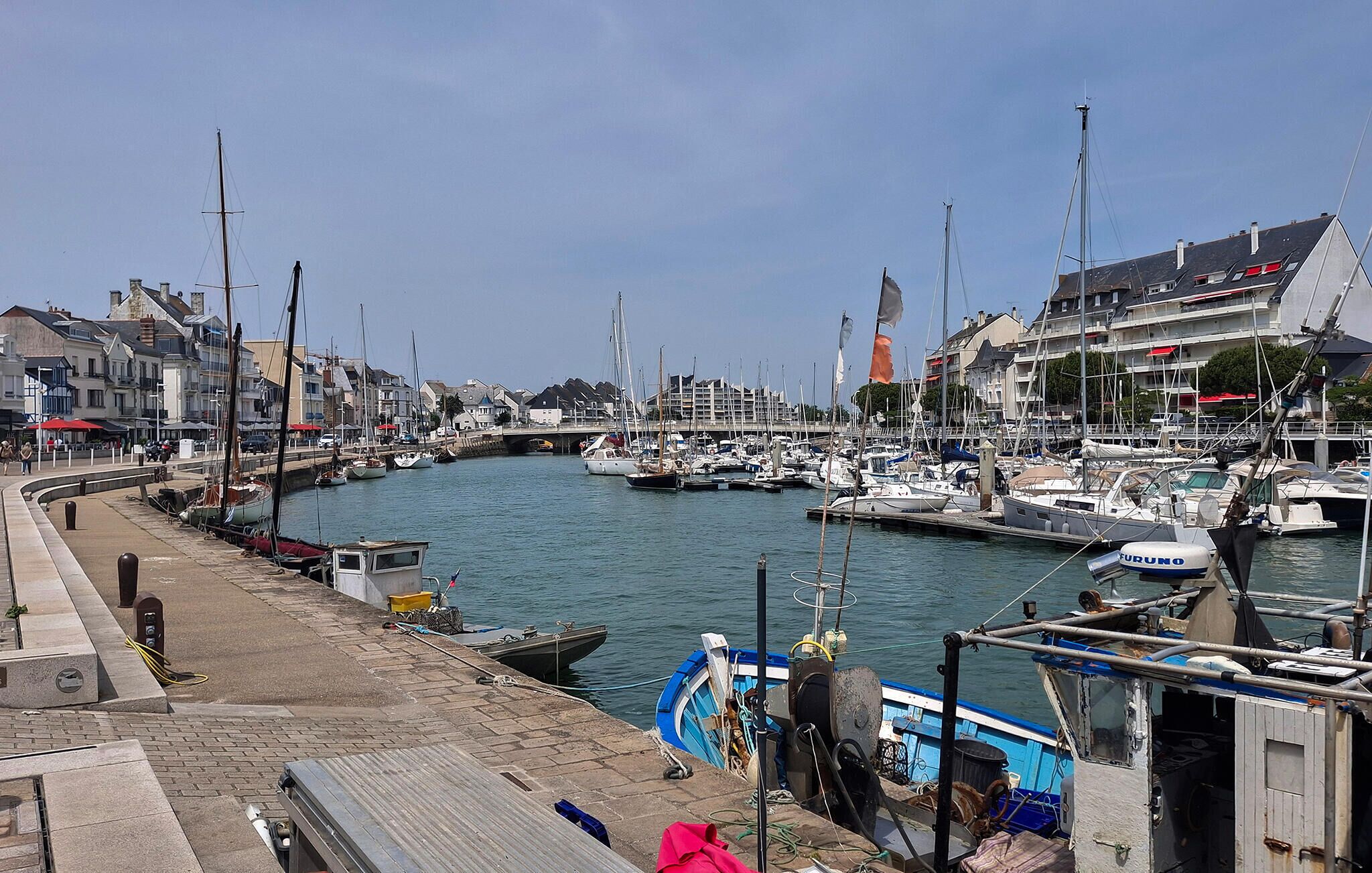 A marina with boats docked, a waterfront promenade, and buildings along the shore.