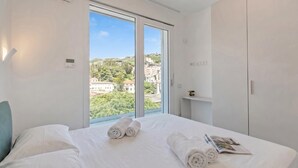 Habitación de hotel con un ventanal grande, una cama con ropa de cama blanca y vistas a edificios y zonas verdes.