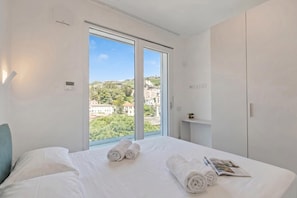 Ein Hotelzimmer mit großem Fenster, einem Bett mit weißer Bettwäsche und Blick auf Gebäude und Grünflächen.