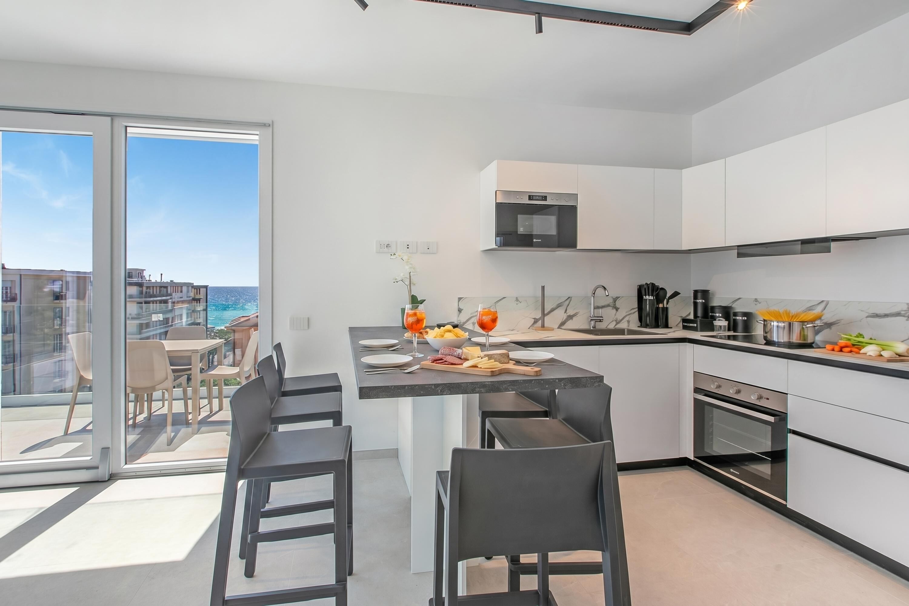 Una cucina moderna con isola centrale, zona pranzo con sedie e vista sul mare attraverso ampie finestre.