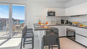 Een moderne keuken met een centraal eiland, een eethoek met stoelen, en uitzicht op zee door grote ramen.