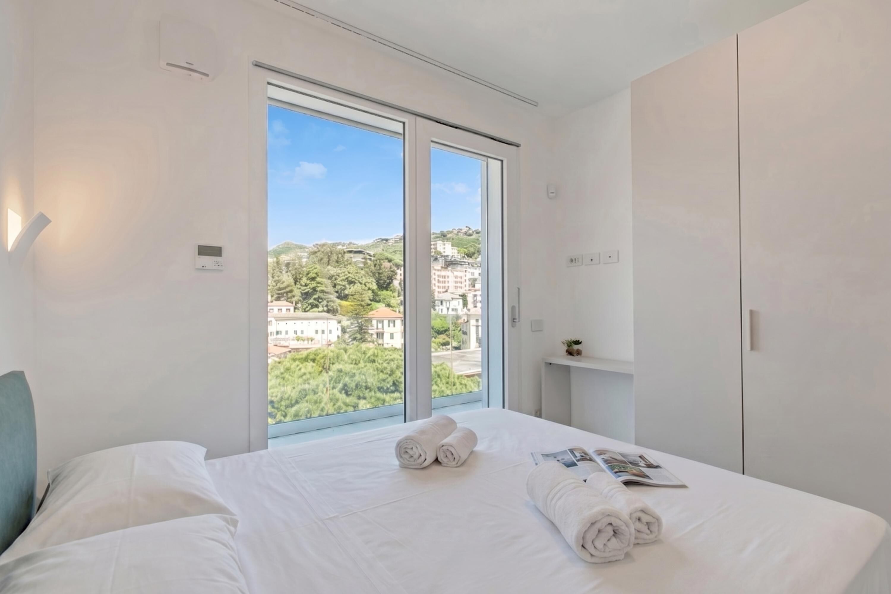 Una camera d'albergo con una grande finestra, un letto con lenzuola bianche, un comodino e un armadio.