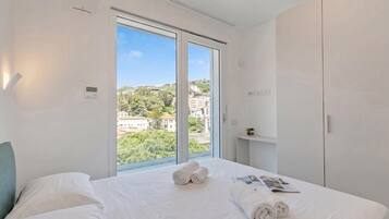 Una camera d'albergo con una grande finestra, un letto con lenzuola bianche, un comodino e un armadio.