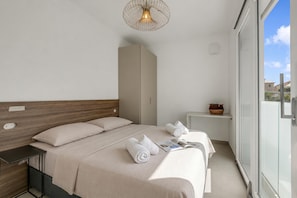 Ett modernt hotellrum med en säng, sängbord, en liten hylla med en skål och ett stort fönster.