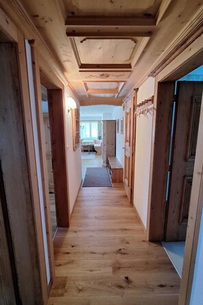 Corredor de madeira com um banco, uma porta e vista para um quarto com mesa e cadeiras.
