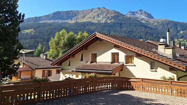 Un tradizionale chalet con tetto in tegole, balcone in legno e vista su una catena montuosa.
