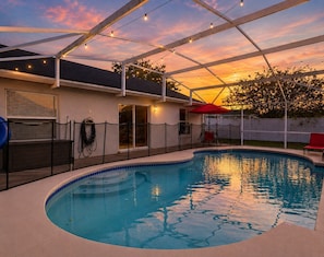 Ein Schwimmbecken mit überdachter Terrasse, einem roten Sonnenschirm und Blick auf den Sonnenuntergang.