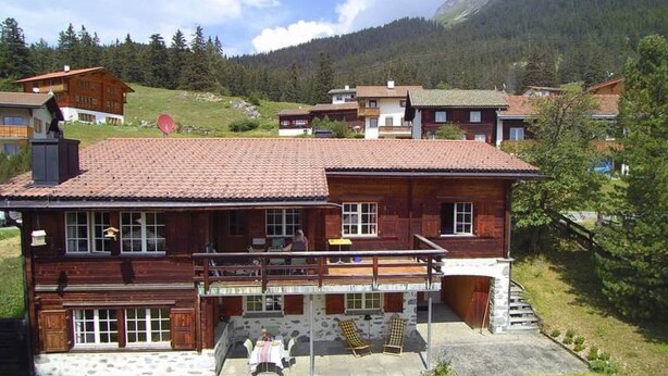 A wooden chalet with a balcony, stone foundation, and a view of the surrounding mountains.