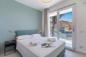 A hotel room with a bed, a nightstand, a reading lamp, a window with a city view, and a towel on the bed. - Nua Boutique Apartaments 13 Towncentre, Finale Ligure, Italy (Finale Ligure)