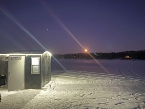 A small, single-story building with a door labeled 'MDNR' and a window, situated in a snowy landscape with a clear sky and a visible moon. - The Narrows on Lawrence Lake
Privacy, Hot Tub, Beach, Dock, Quick Trail Access (Outing)