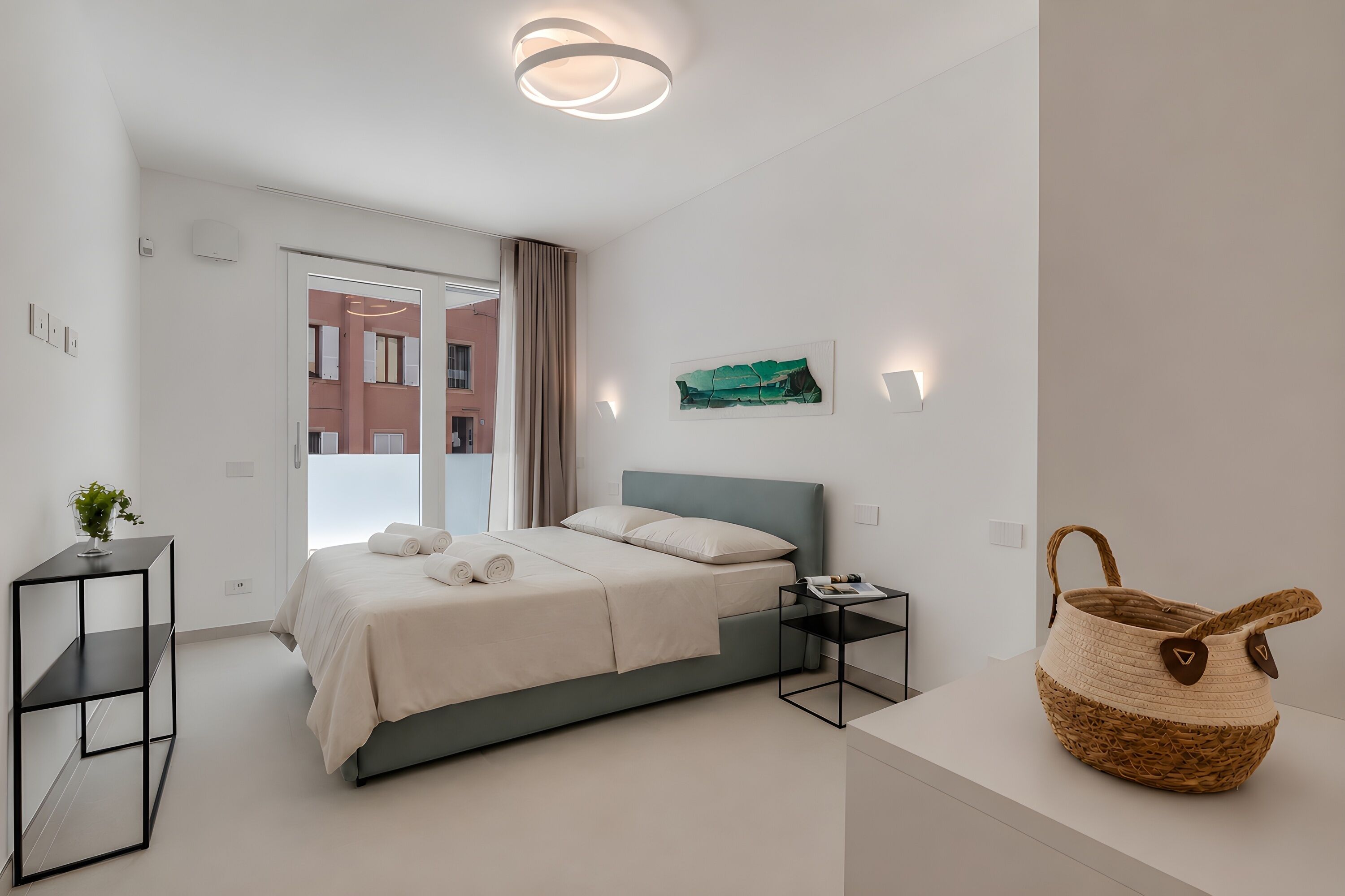 A modern bedroom with a bed, bedside tables, a wall-mounted light, and a view of a building outside.