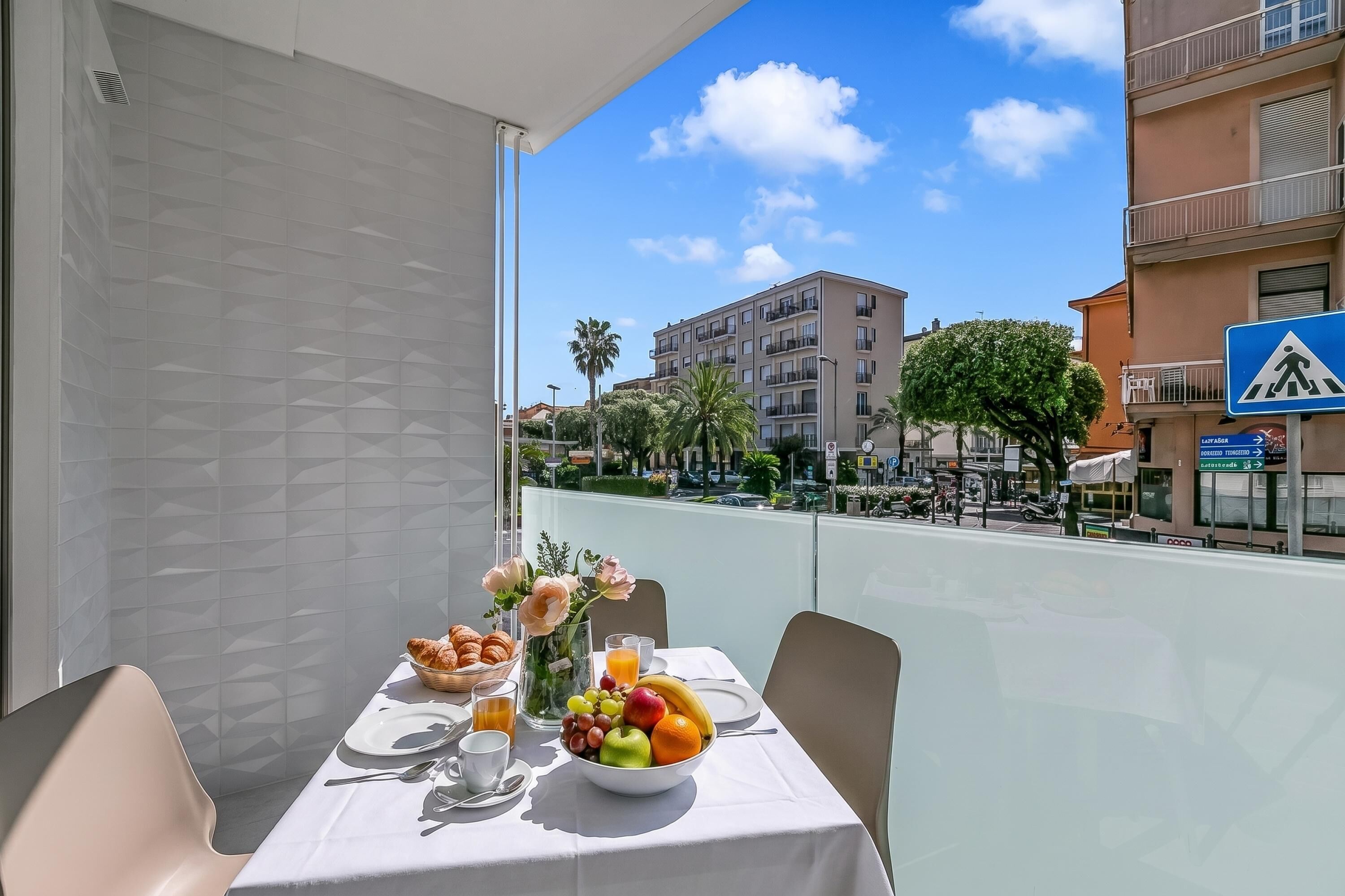A balcony with a table set for breakfast, a bowl of fruit, a vase with flowers, and a view of the cityscape.