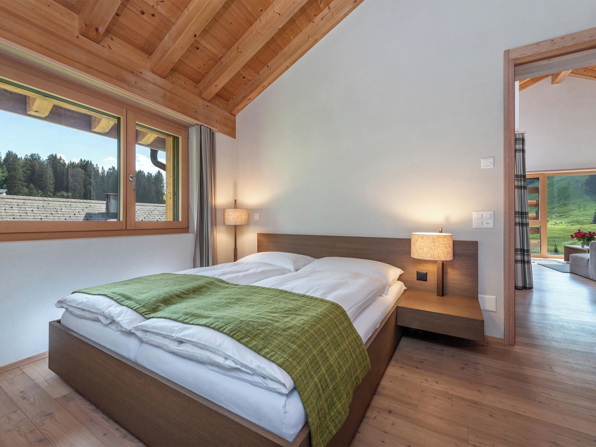 Un dormitorio con una cama de madera, dos lámparas de noche, vistas a un bosque por una ventana y una puerta que da a otra habitación.