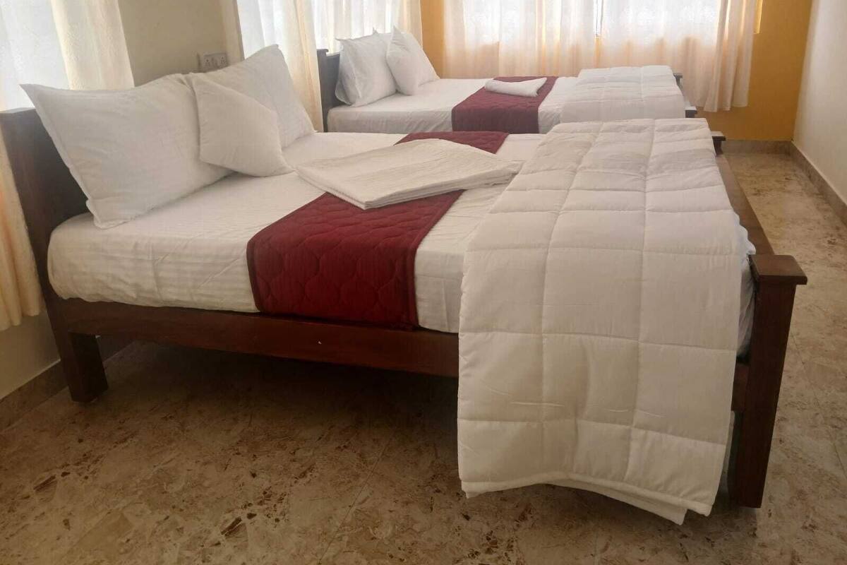 Habitación de hotel con dos camas, cada una con ropa de cama blanca y una manta roja. El piso es de baldosas y hay una ventana con cortinas traslúcidas.