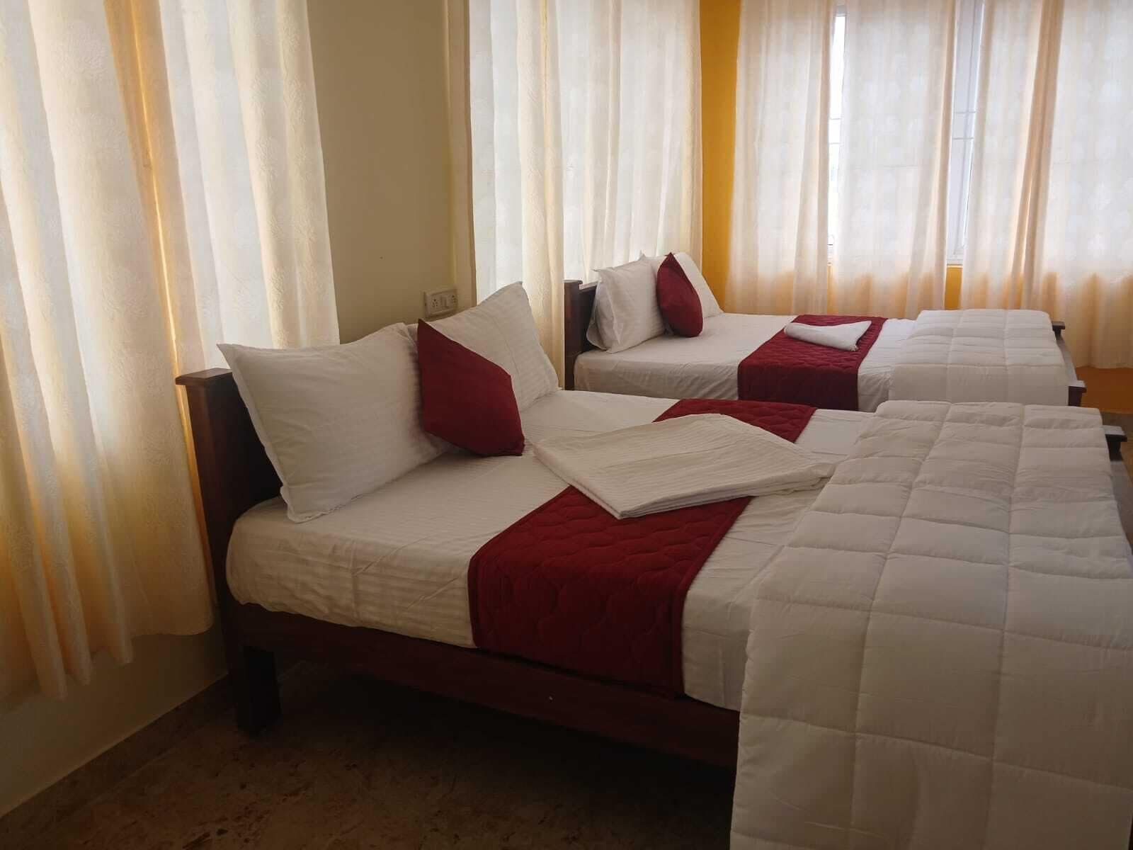 Habitación de hotel con dos camas, cada una con ropa blanca y detalles rojos, y un ventanal con cortinas traslúcidas.
