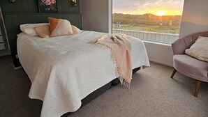 A bedroom with a large bed, a view of the sunset, a chair, and a wall-mounted light fixture. - Stunning 3-bedroom house with the best beach/ocean views (Foxton Beach)
