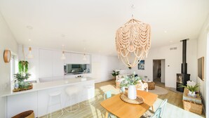 A modern kitchen with white cabinetry, a central island, bar stools, a dining table with chairs, and a hanging light fixture. - Stunning 3-bedroom house with the best beach/ocean views (Foxton Beach)
