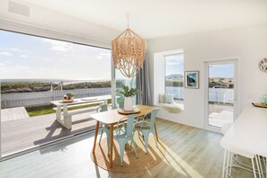 Et rummeligt værelse med et stort vindue, et træbord med stole, en hængende lampe og udsigt over landskabet.