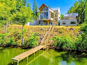 Une maison moderne dotée d’une terrasse en bois qui mène à un quai surplombant l’eau, le tout entouré d’arbres.