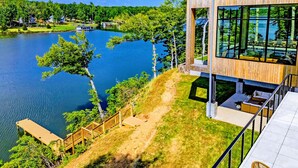 Ein modernes Gebäude mit großen Fenstern, die auf einen See blicken, einer Holzterrasse und einem wolkenlosen Himmel.