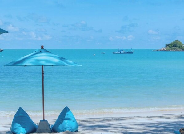 Una platja amb un parasol blau i dos coixins blaus, mar cerúlle i un vaixell a la distància.