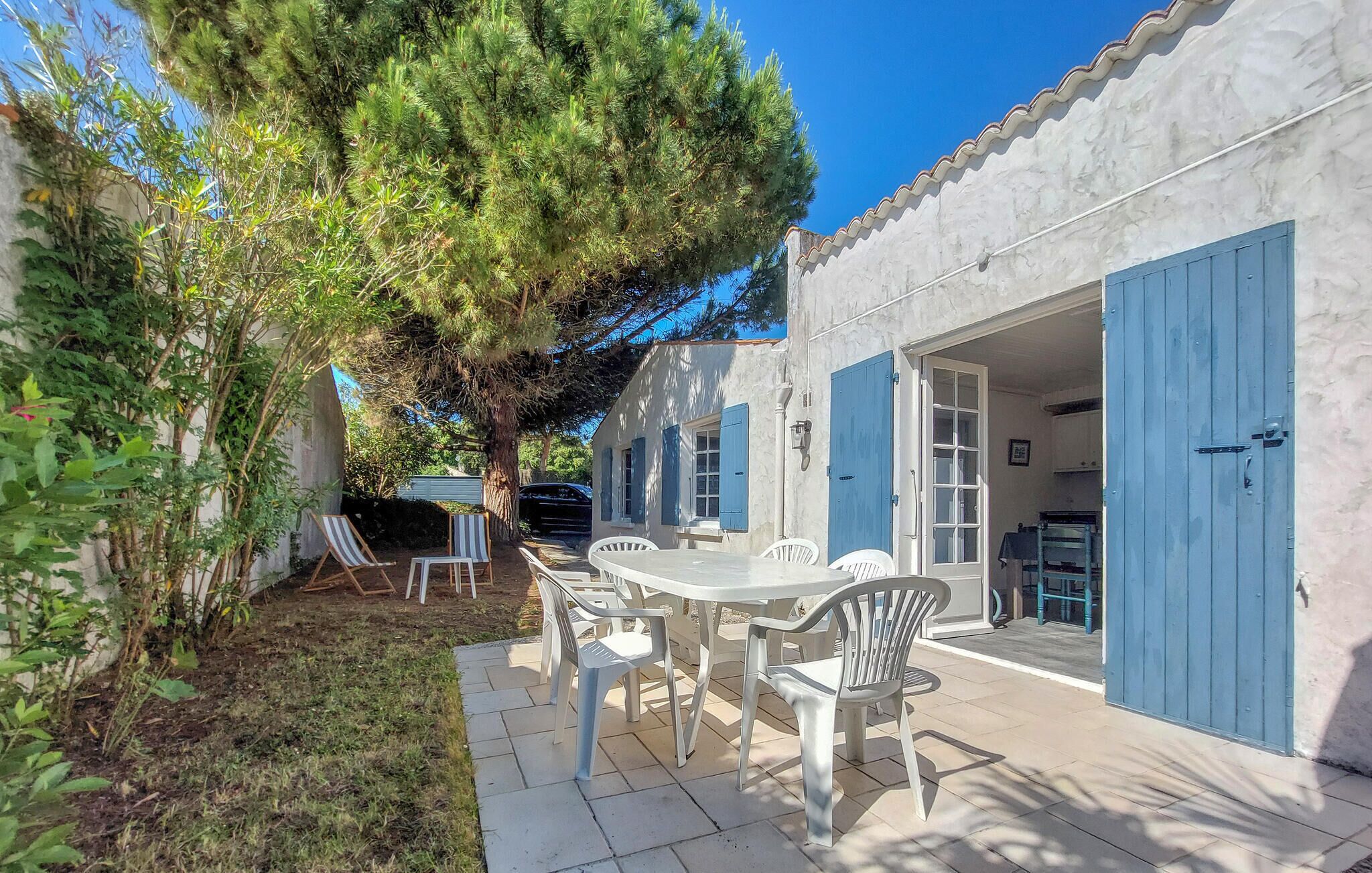 Un patio con una mesa, sillas, un árbol y un edificio con contraventanas azules.