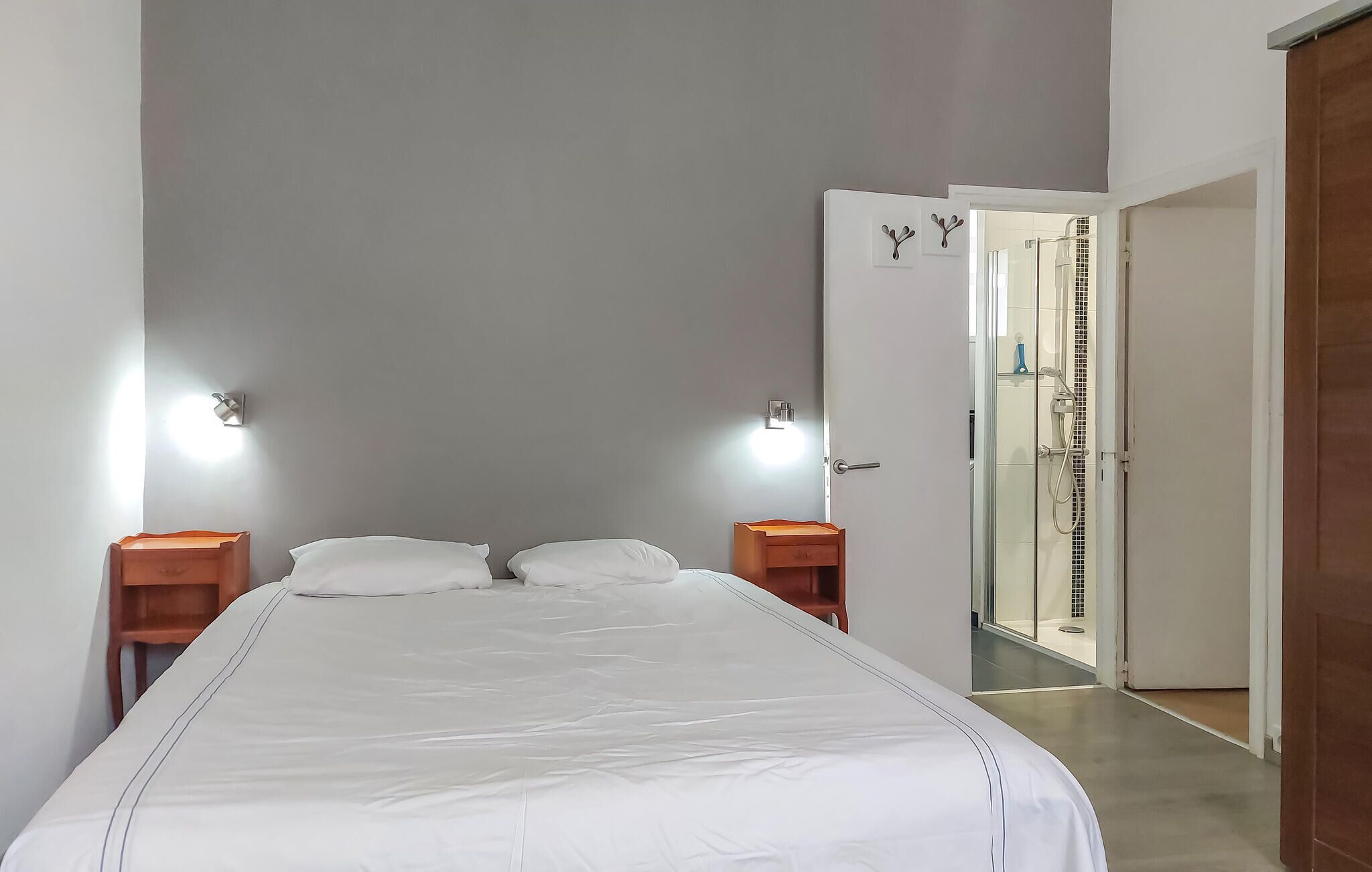 A bedroom with a bed, two bedside tables, a nightstand, a wall-mounted light, and a door leading to a bathroom.