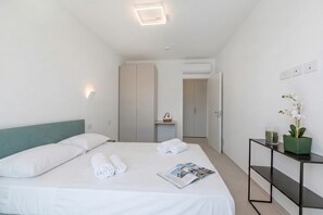Ein modernes Hotelzimmer mit einem ordentlich bezogenen Bett, einem Nachttisch mit einer Zeitschrift, einer kleinen Pflanze und einer Wand-Klimaanlage.