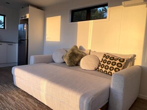 A modern living room with a sofa, pillows, and a built-in kitchen area. - The Cabin & Sauna @ The Highlands (Waipu)