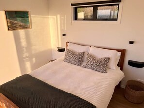 A bed with a white bedspread and two patterned pillows, a window above, and a wall-mounted shelf. - The Cabin & Sauna @ The Highlands (Waipu)