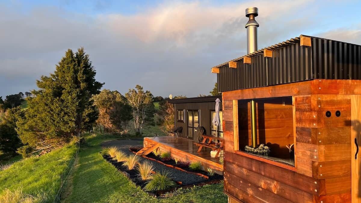 Une cabane moderne en bois, dotée d’une terrasse, entourée d’un jardin aménagé et d’arbres.