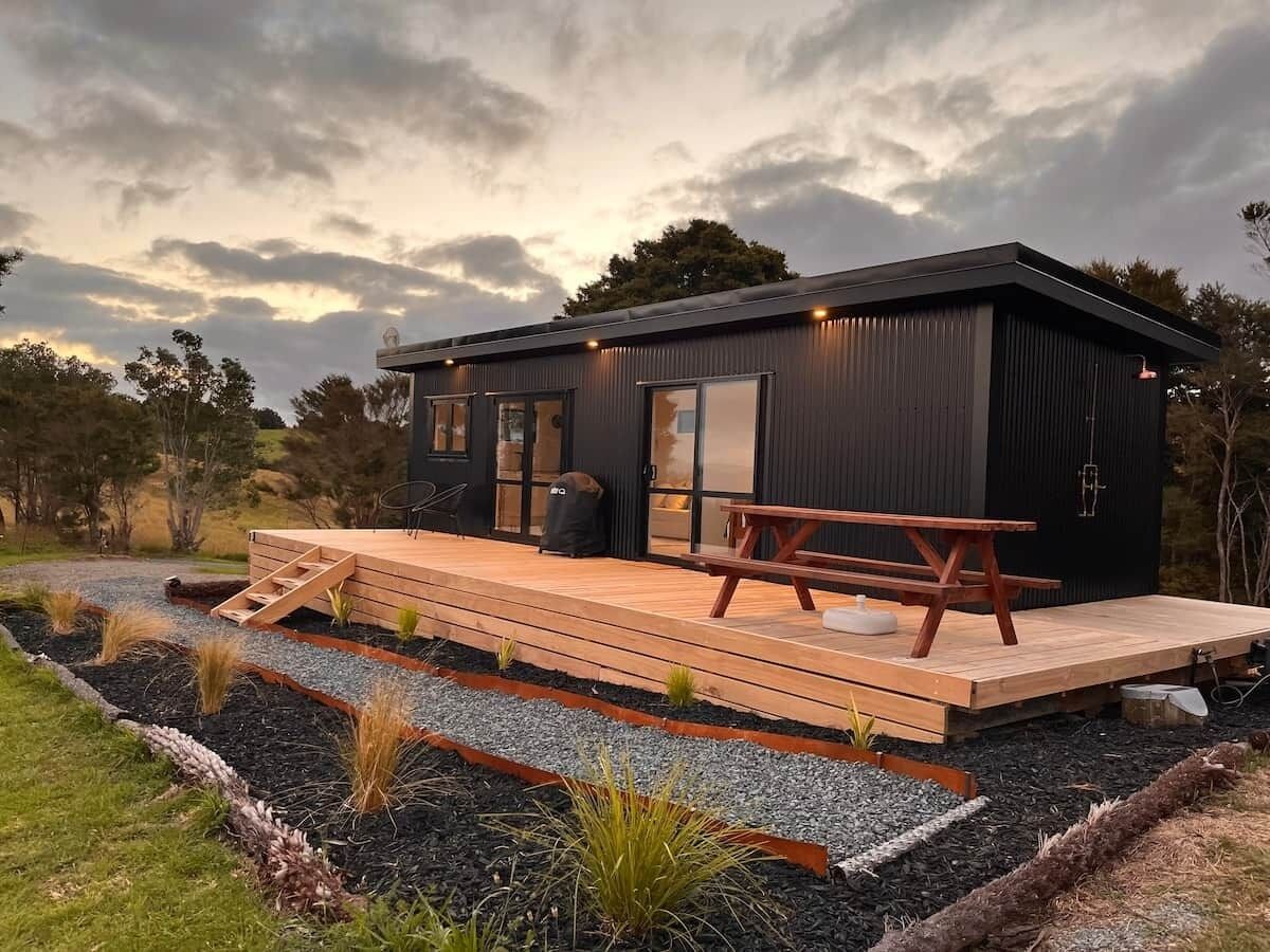 A modern cabin with a wooden deck, a picnic table, and a black exterior.