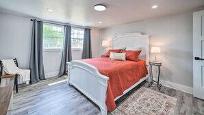 Une chambre avec un lit blanc, des rideaux gris, une petite chaise, une table de chevet avec une lampe et un tapis à motifs.