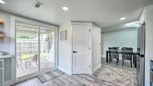 Une pièce dotée d’une porte-fenêtre coulissante donnant sur un jardin, d’un coin repas avec une table et des chaises, et d’un espace cuisine avec un réfrigérateur.