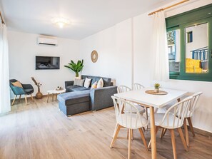 A modern living room with a grey sectional sofa, a white dining table with chairs, a TV mounted on the wall, and a large window with a view of a building. - Gemini Villas- Harmony Villa by Interhome (Rhodos City)