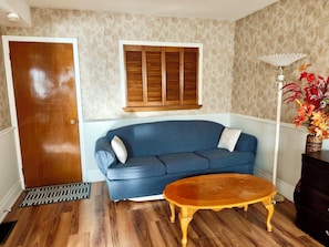 Un salon avec une porte en bois, un canapé bleu, une table basse en bois, une fenêtre avec des volets en bois et un lampadaire agrémenté d’un arrangement floral.