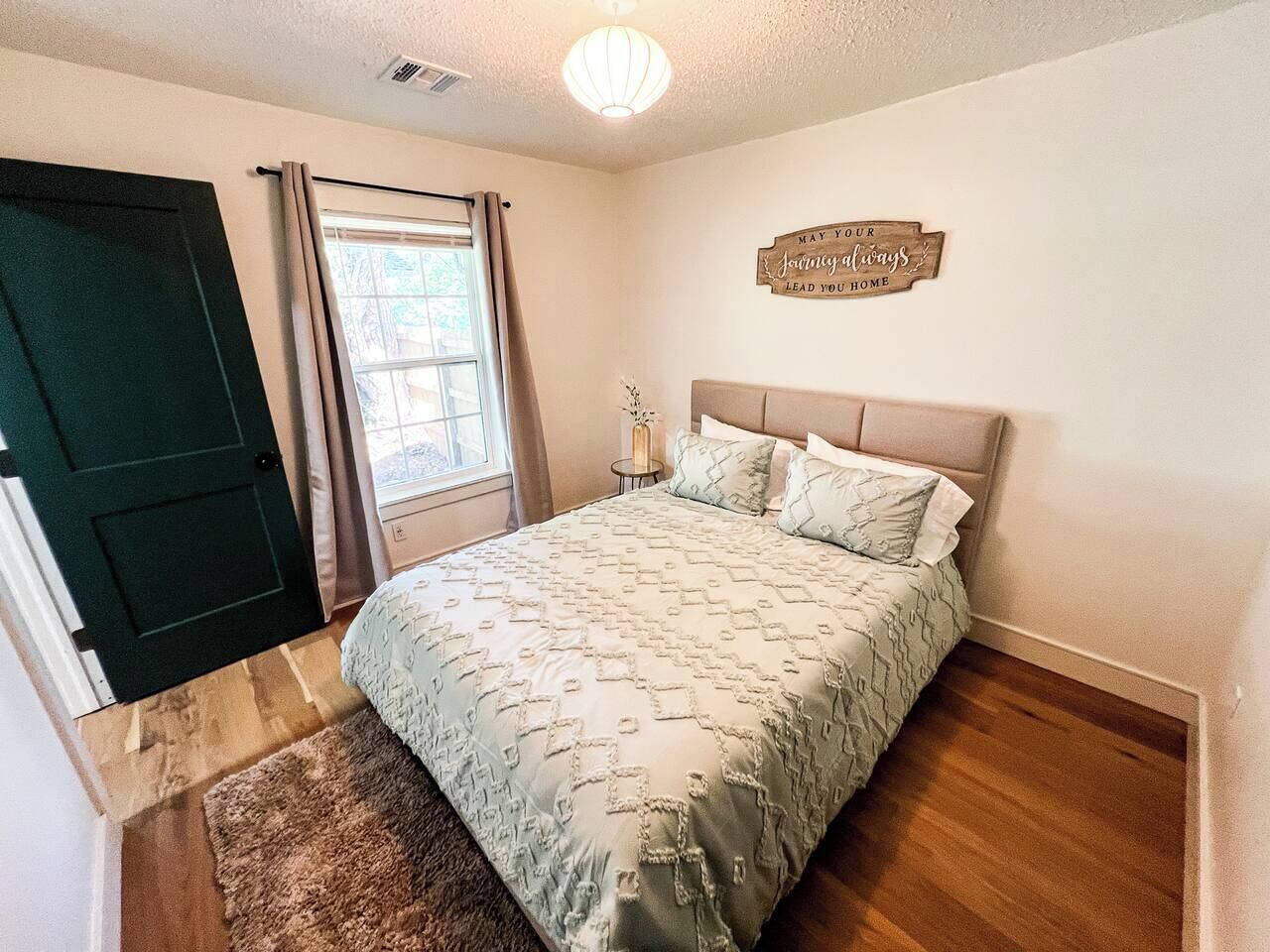A bedroom with a bed, a door, a window with curtains, a bedside table, and a wall sign.