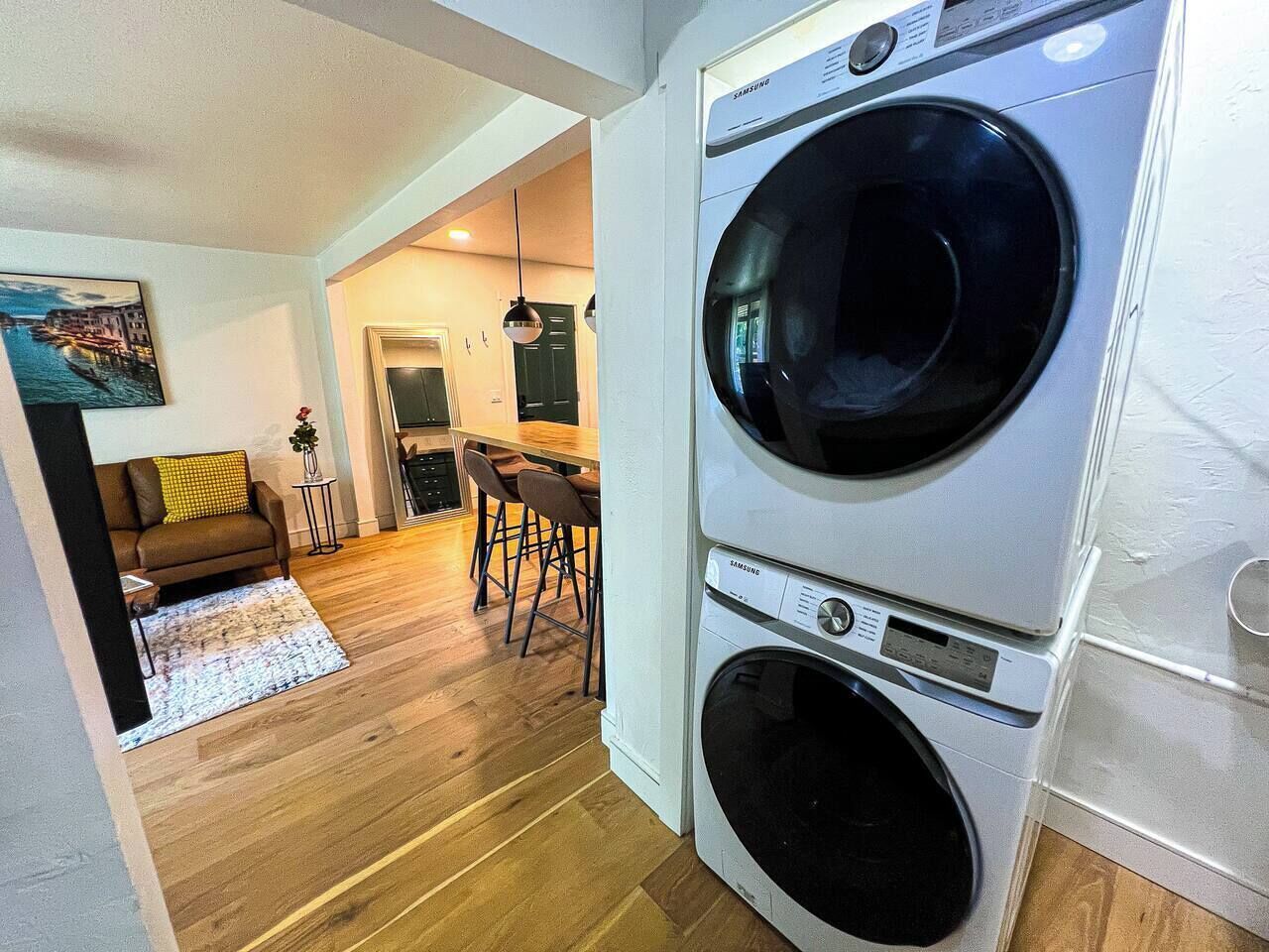 Une buanderie équipée d’un lave-linge et d’un sèche-linge Samsung, un coin repas avec des chaises et une table, et un salon avec un canapé et un tableau.