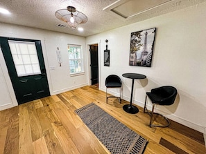 Ein Zimmer mit Holzboden, einem schwarzen Teppich, zwei schwarzen Stühlen, einem kleinen runden Tisch, einer schwarzen Wanddekoration und einer Tür mit Fenster.