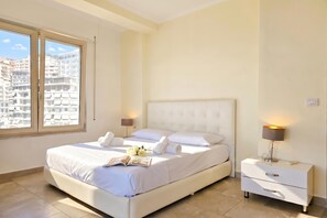 A hotel room with a bed, bedside lamps, a nightstand, and a window with a view of buildings. - Happy Stay Vincent - 270 /w Sea View Balcony, Sarandë, Albania (Sarandë)