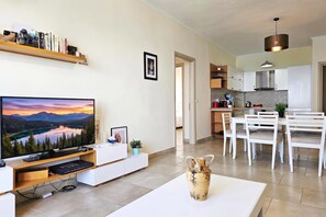 A modern living room with a flat-screen TV, a white coffee table, and a dining area with white chairs. - Happy Stay Vincent - 270 /w Sea View Balcony, Sarandë, Albania (Sarandë)