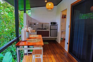 A wooden deck with a table and bench, a kitchen with modern appliances, and a view of lush greenery. - Tree House La Fortuna w/Jacuzzi (La Fortuna)