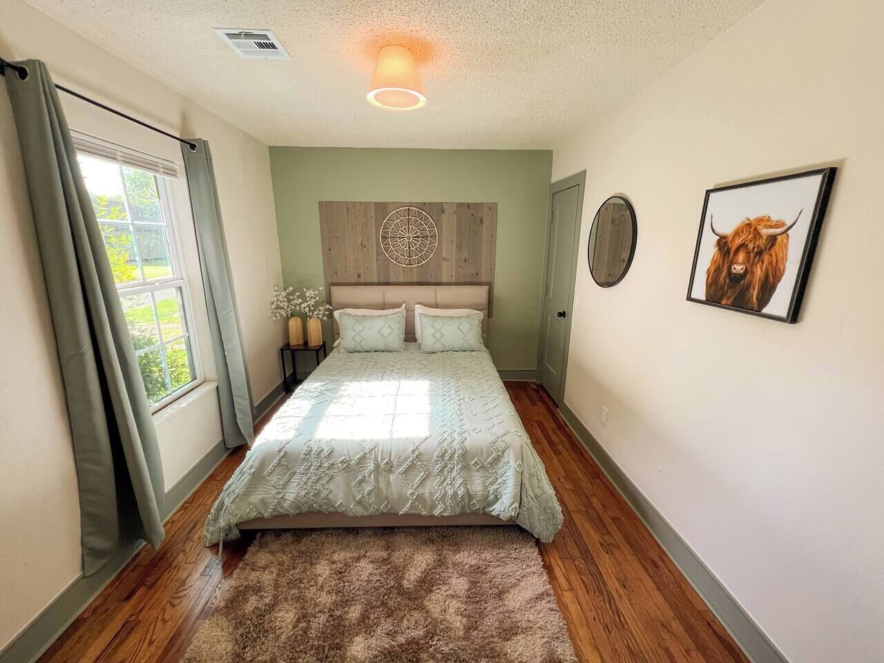 A bedroom with a bed, a window with curtains, a wall-mounted artwork, and a wooden floor.