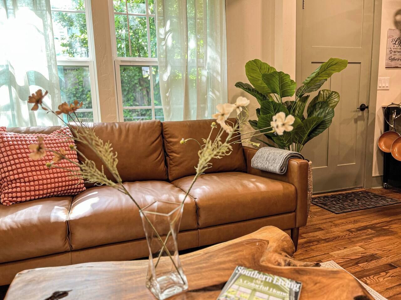 Un canapé en cuir marron, agrémenté d’un vase en verre et d’un magazine, ainsi qu’une grande plante verte et un coussin rouge à motifs.