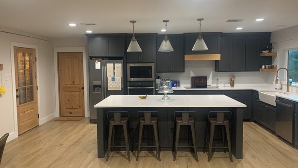 Cozinha moderna com ilha central, banquetas, armários escuros, eletrodomésticos de aço inoxidável e iluminação pendente.