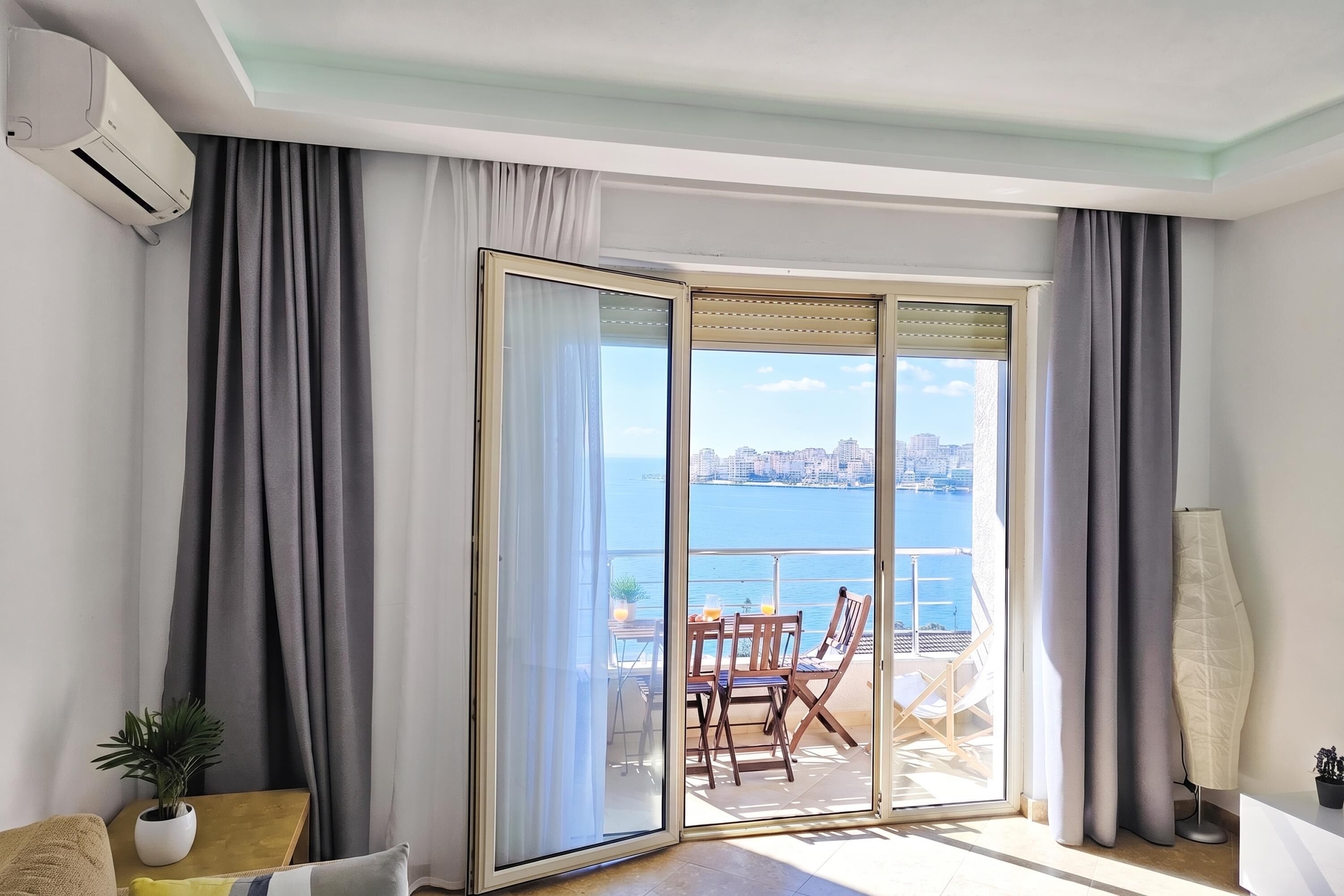 A room with a sliding glass door leading to an outdoor seating area with a view of the sea and cityscape.